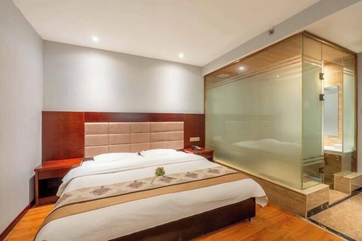 Hotelkamer met een groot bed, een houten hoofdbord, een glazen scheidingswand en een klein nachtkastje.