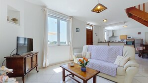 Un salon avec un canapé, un téléviseur posé sur un meuble, une table avec un vase, et une cuisine à l’arrière-plan.