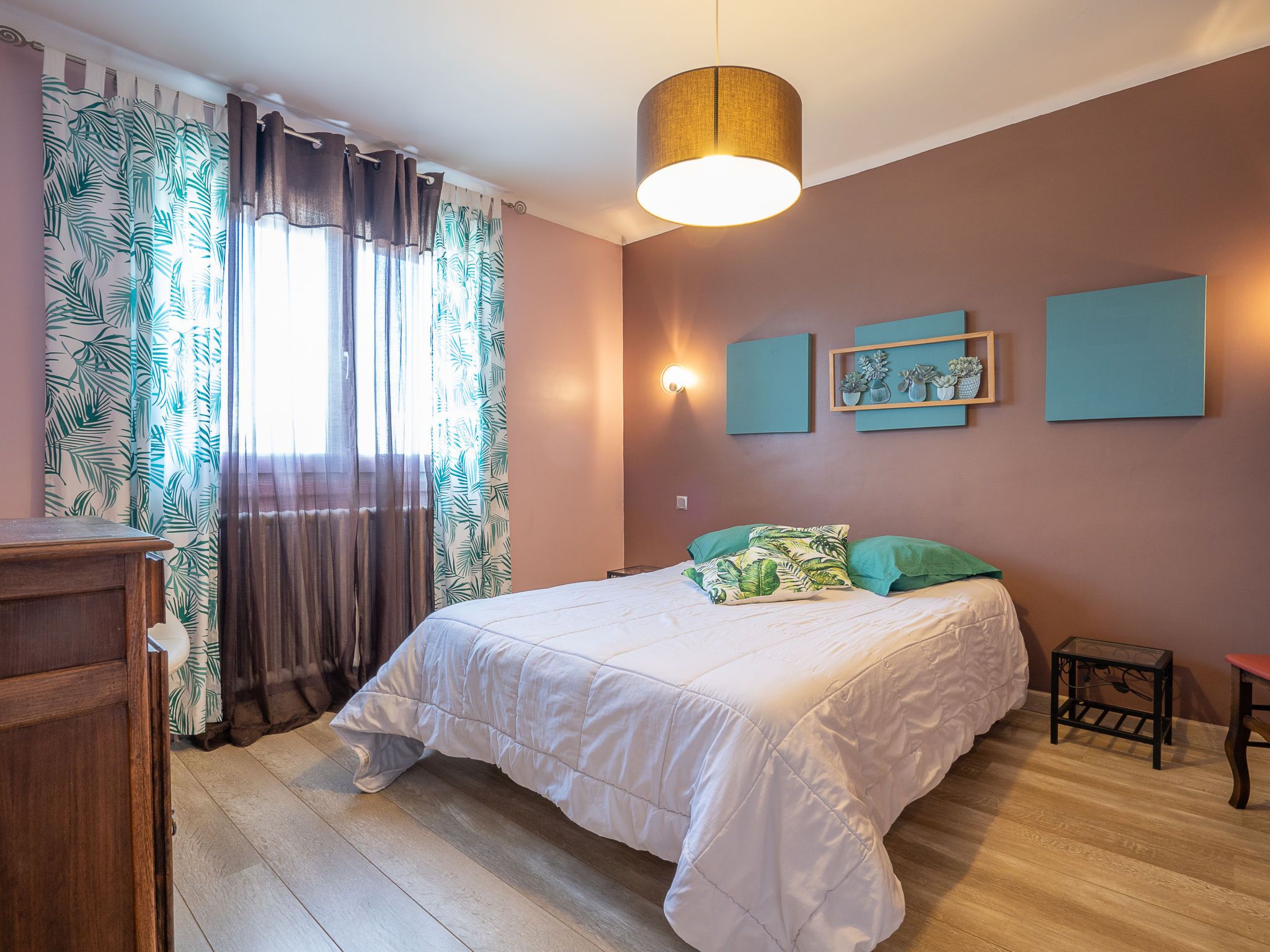Une chambre à coucher avec un lit, des rideaux, une lampe fixée au mur et des tableaux encadrés.
