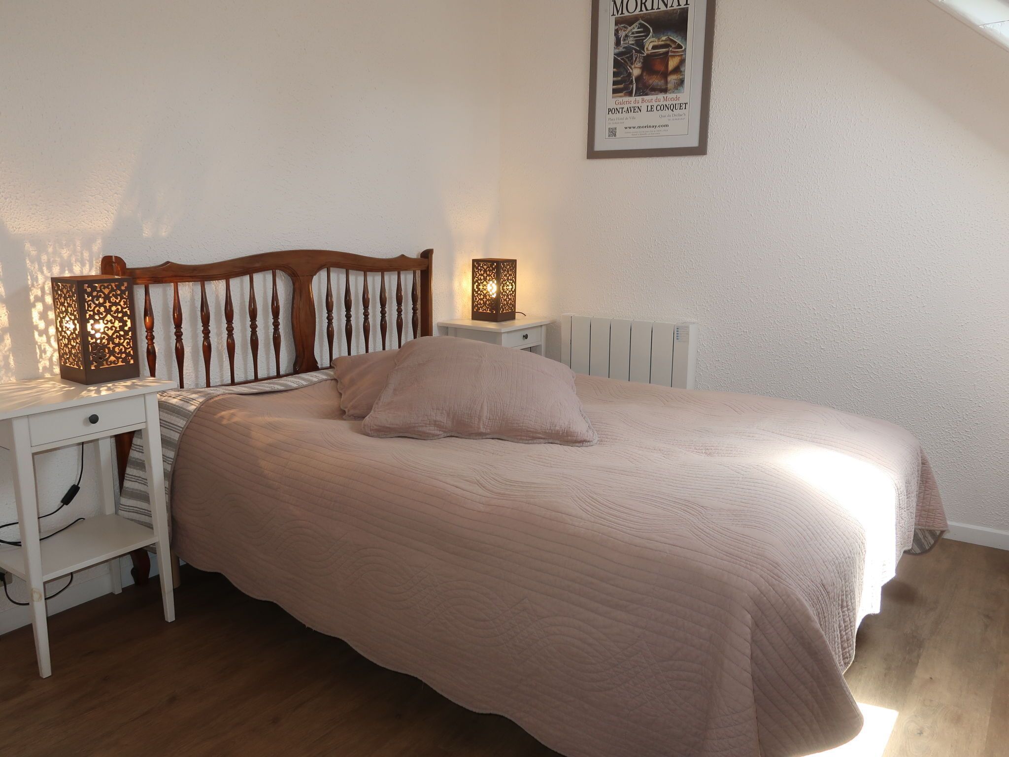 Un dormitorio con cabecera de madera, una mesita de noche con lámpara, cama con manta rosa y un cartel en la pared.