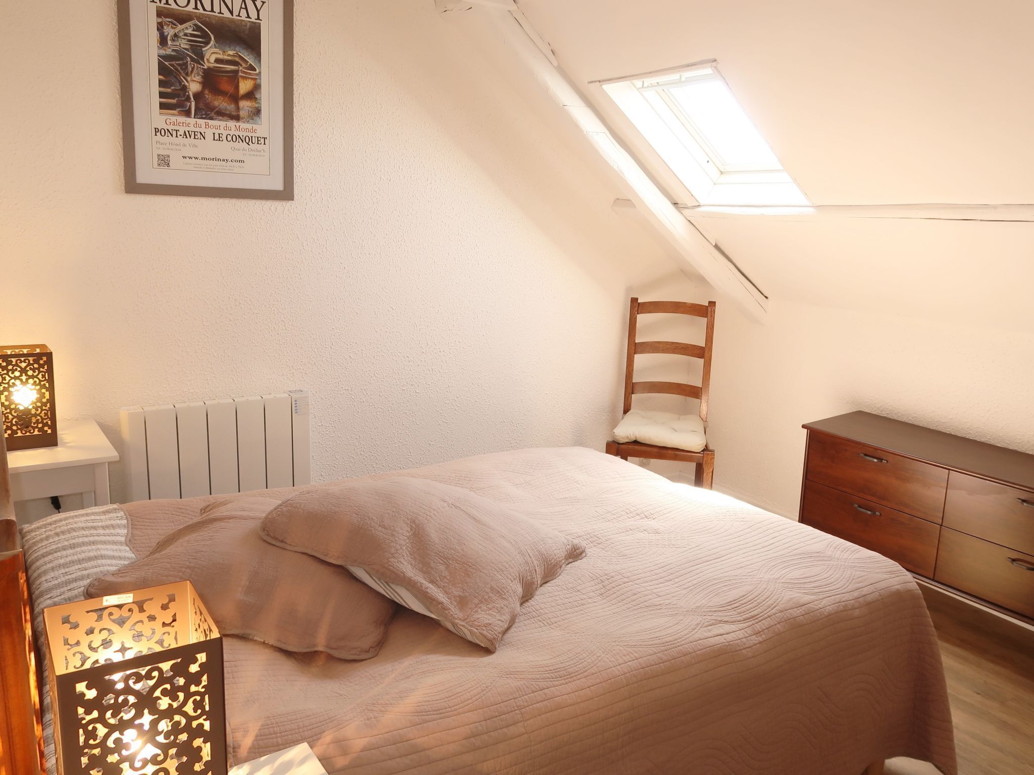 Ett sovrum med ett sluttande tak, ett takfönster, en säng med ett beige överkast, en trästol, ett sängbord med en lampa och en byrå.