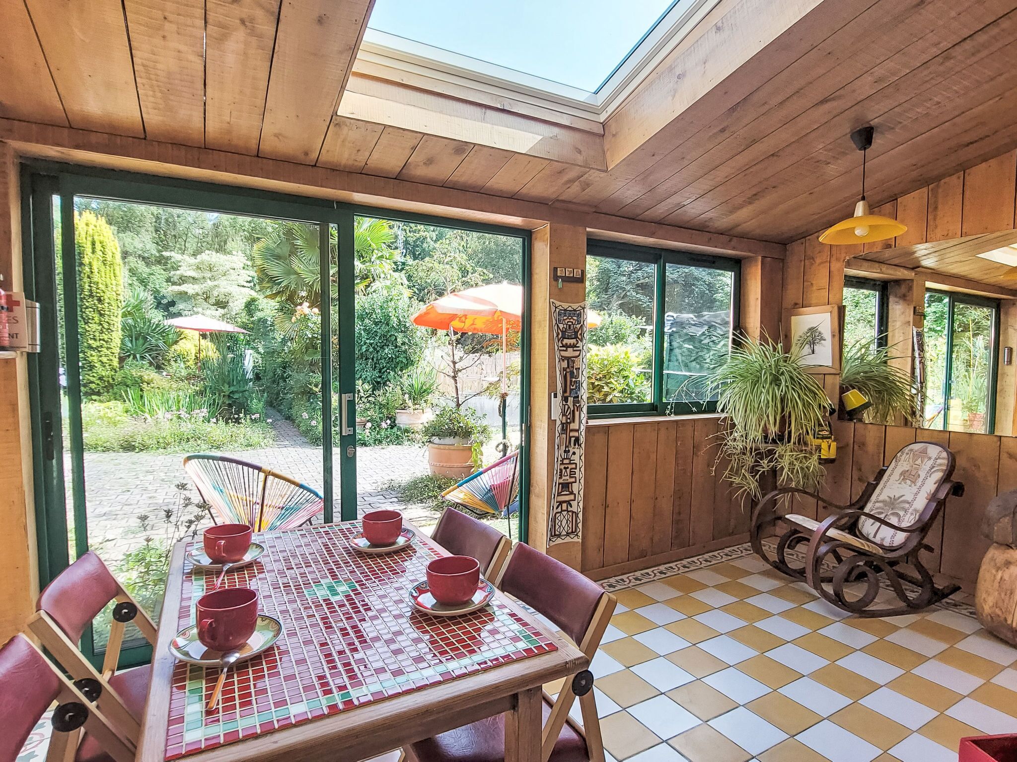 A wooden interior with a checkered floor, a table with red cups, and a view of a garden through large windows.