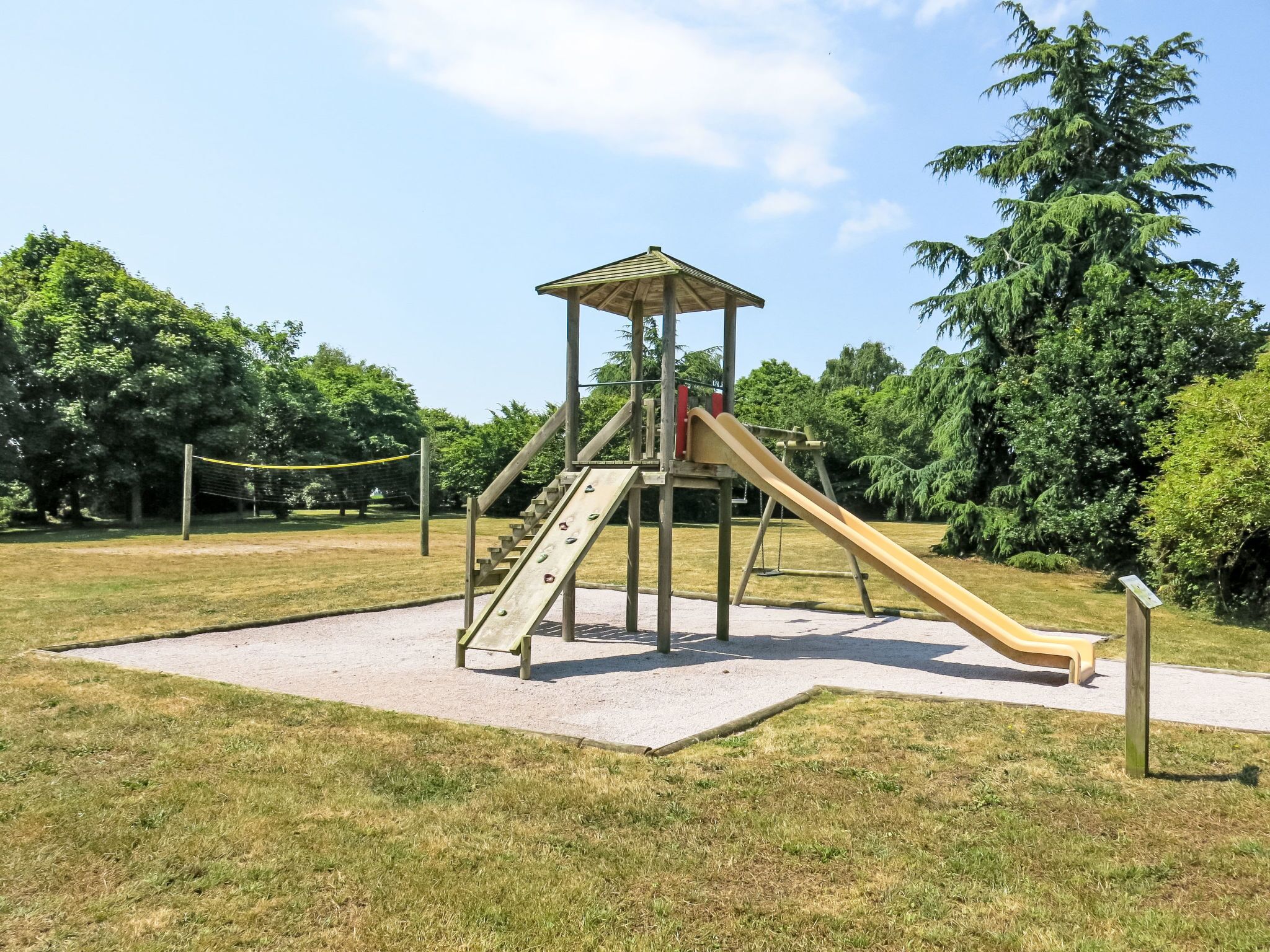 A playground with a slide and climbing area, surrounded by grass and trees.