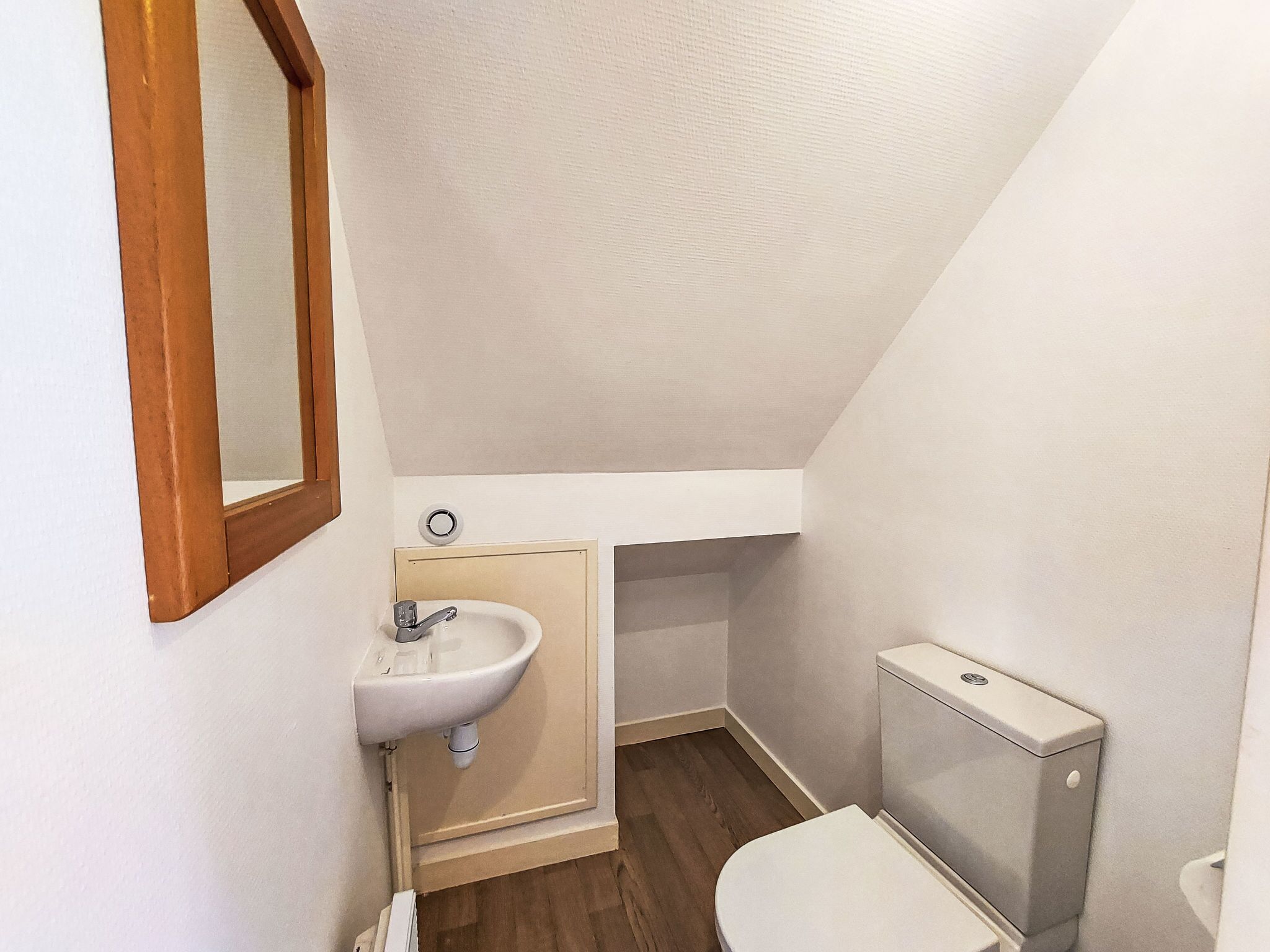 A small, compact bathroom with a wooden-framed mirror, a white sink, and a toilet.