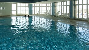 Una piscina interior con agua cristalina, rodeada de ventanas y sillas blancas.