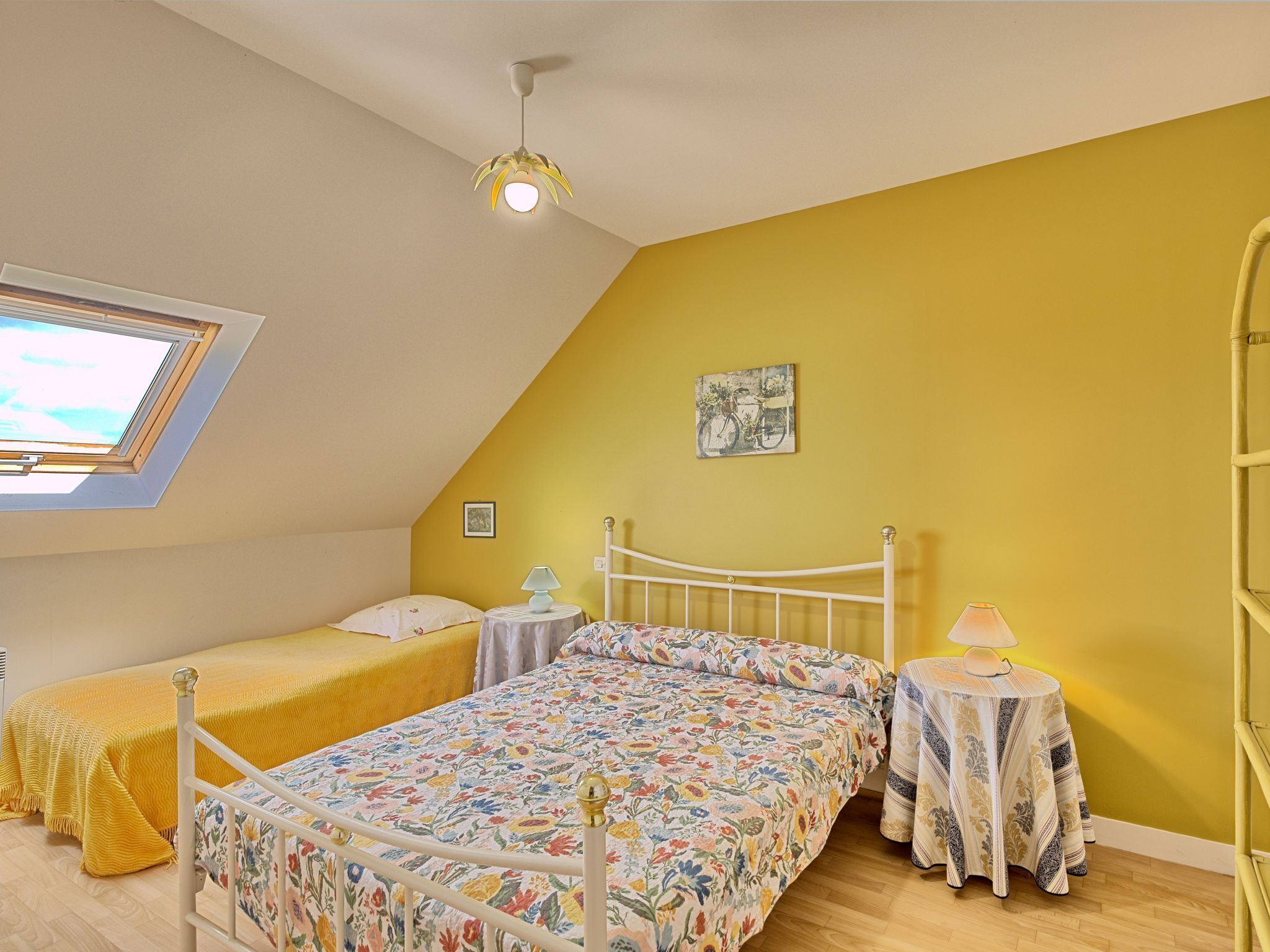 Ein Schlafzimmer mit geneigter Decke, einem Oberlicht, einem Bett mit floralem Überwurf, einem Nachttisch mit Lampe und einem Wandbild mit einem Fahrrad.