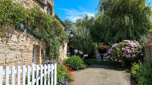 En bygning i sten med et hvidt stakittancehegn, omgivet af frodigt grønt og blomstrende planter.