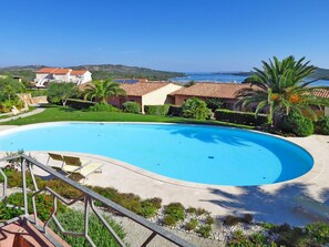 A large swimming pool with a curved edge, surrounded by a landscaped area with palm trees and a view of the sea. - Il Fjordo bilo (PPZ164) by Interhome (Porto Pozzo)