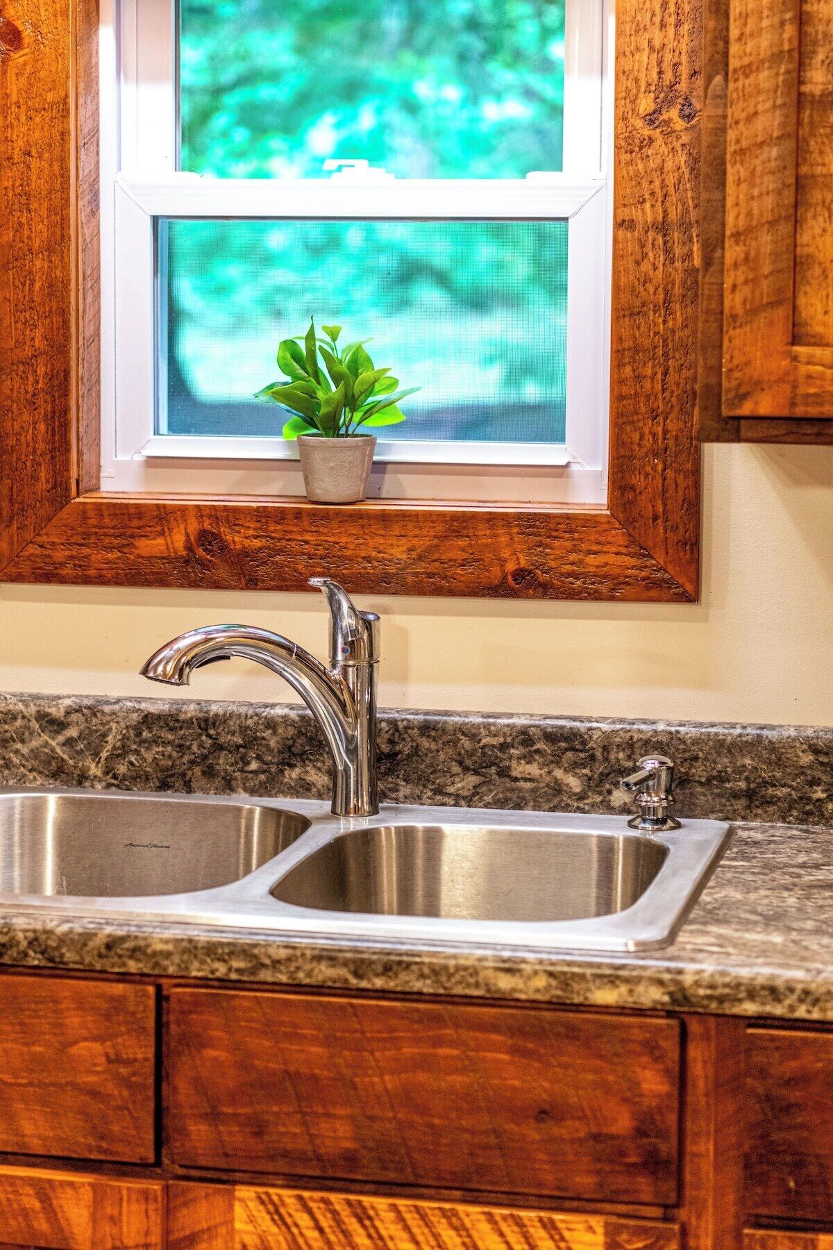 A kitchen with a double stainless steel sink, wooden cabinets, a window with a view of greenery, and a small potted plant on the windowsill.