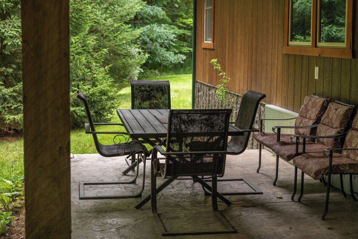 A covered patio with a table and chairs, a bench, and a wooden building in the background.