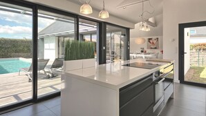 A modern kitchen with a large island, glass doors leading to a pool area, and pendant lighting. - Villa Noalou by Interhome (Arzon)