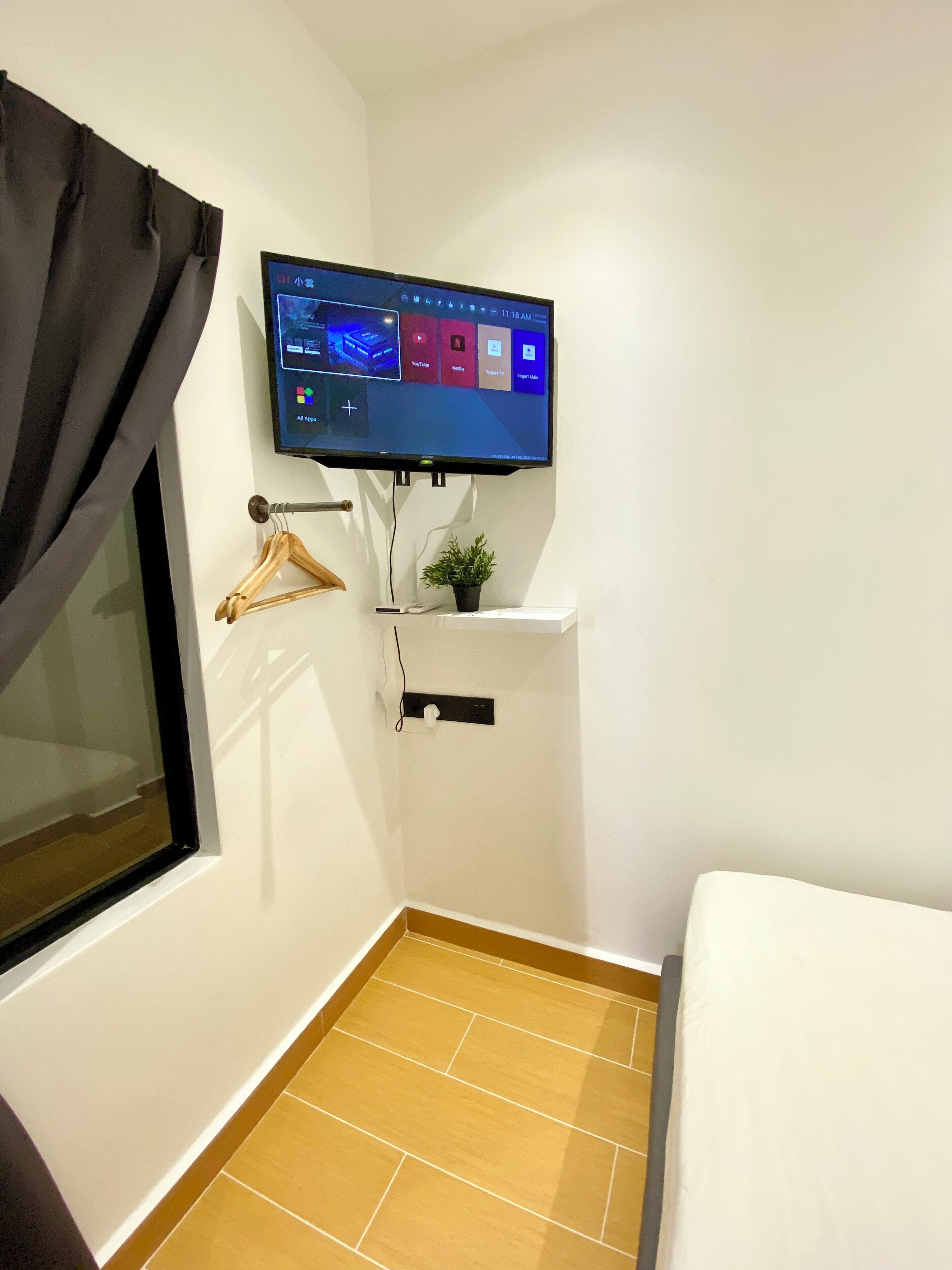 A small room with a TV mounted on the wall displaying various apps, a small shelf with a plant, and a window with a curtain.