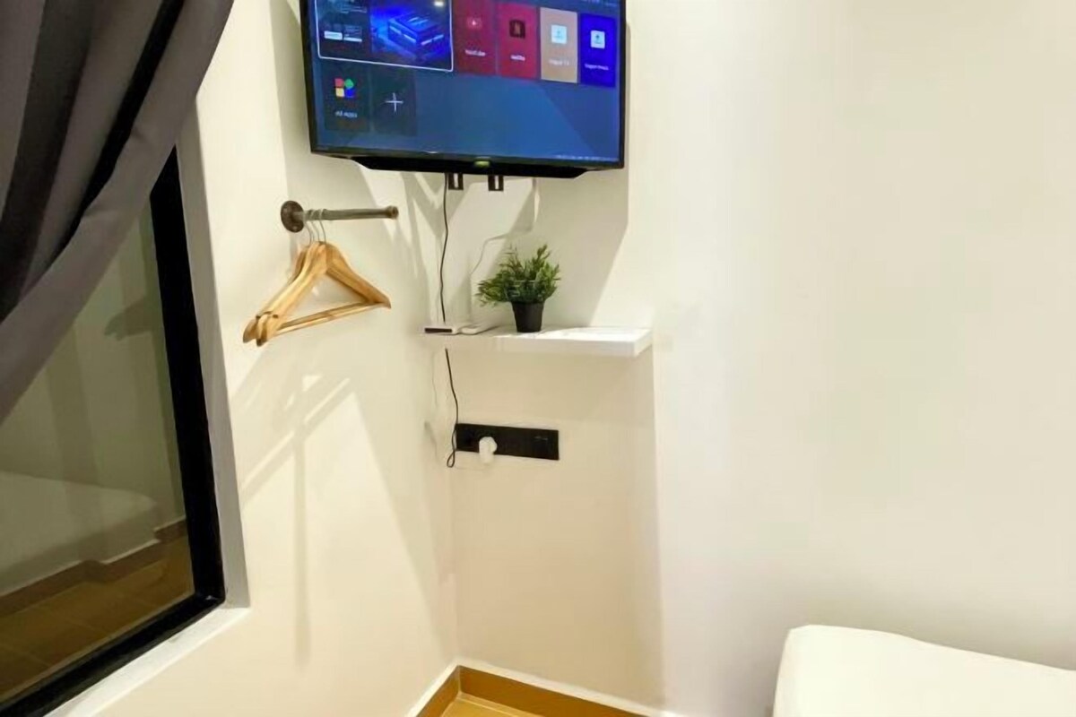 A small, clean room with a window, a mounted TV, a wooden hanger, and a small plant on a white cabinet.