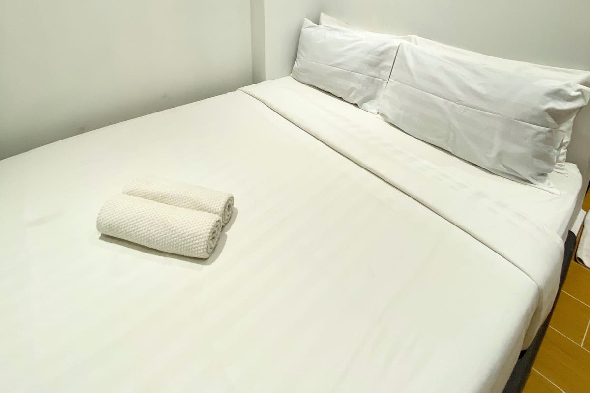 A single bed with white bedding and pillows, a rolled white towel, and a wall-mounted air conditioner.