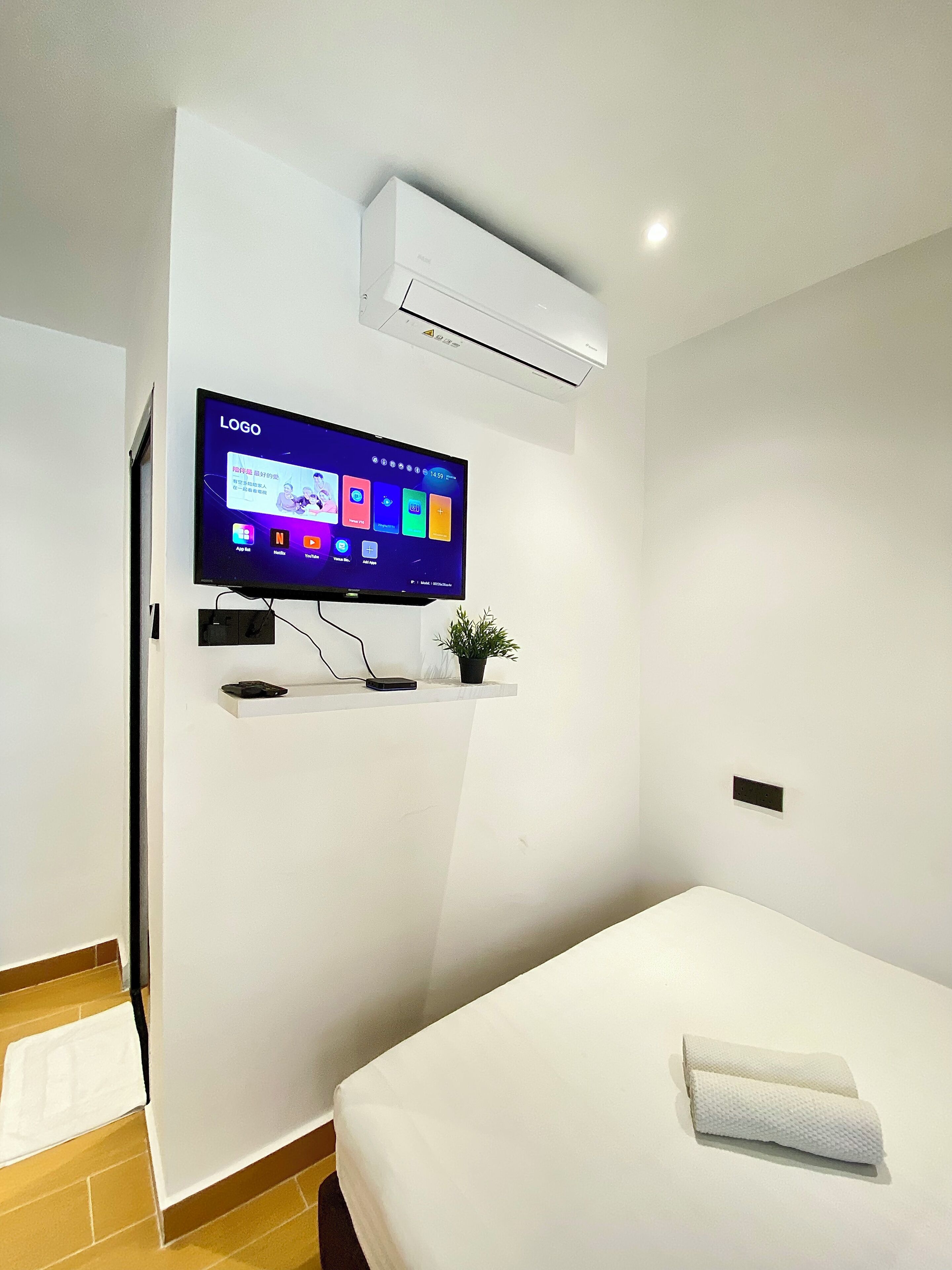 Quarto de hotel moderno com televisão de tela plana na parede, uma pequena estante com uma planta e uma cama bem arrumada com toalha.