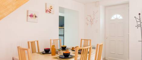 A dining area with a wooden table, six chairs, and a chandelier. There is a white door and a coat rack to the right.