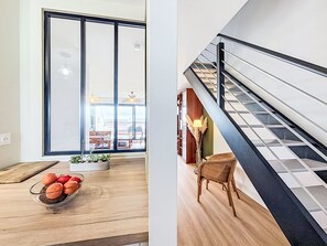 Un interior moderno con un gran ventanal corredero, una mesa de comedor de madera con un frutero y una escalera con barandilla de metal.