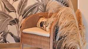 A wicker chair with a cushion, a vase with dried plants, and a wall with a tropical leaf pattern. - Vacation home Hoedic by Interhome (Plouhinec Morbihan)