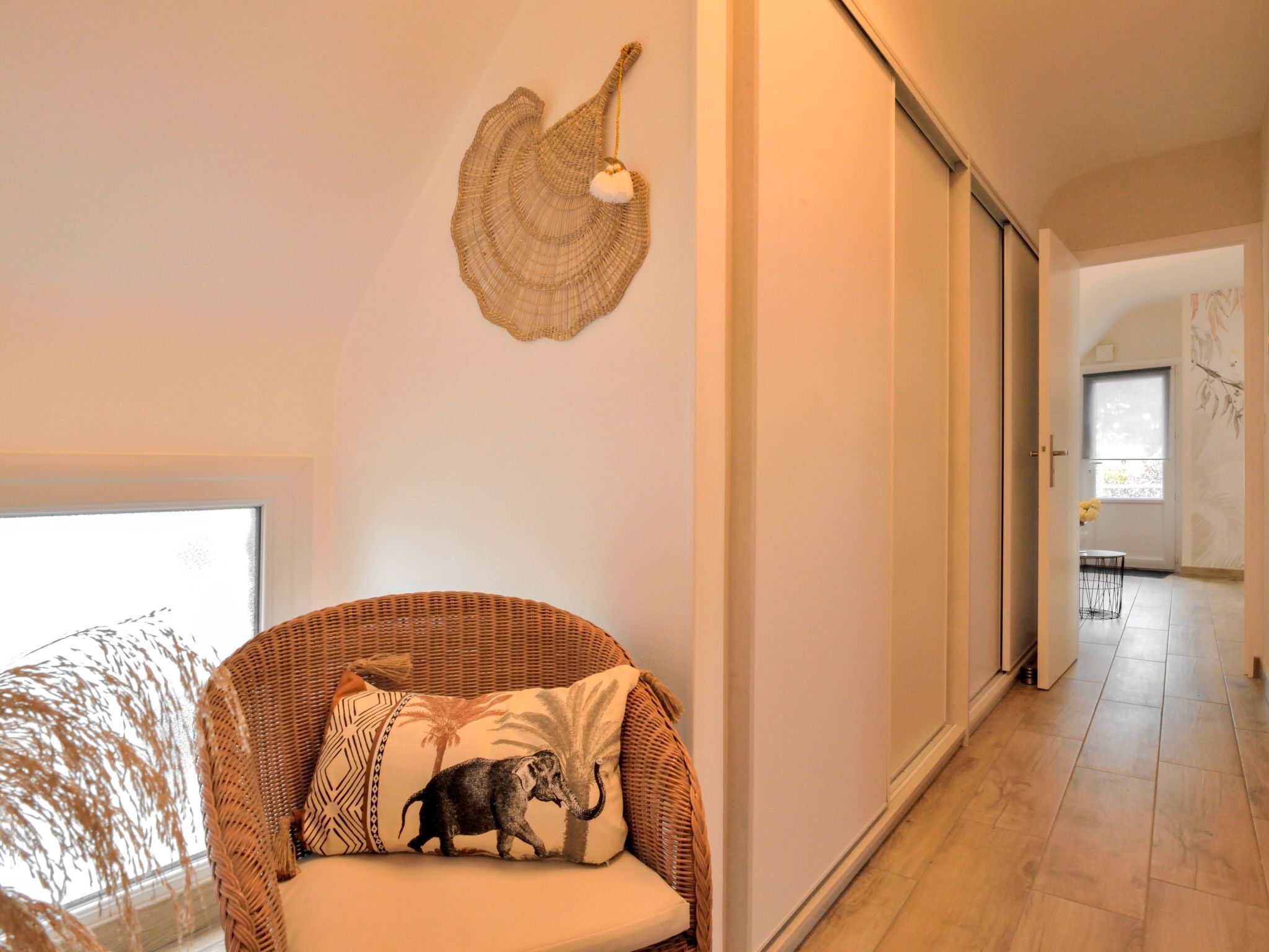 Un interior modern amb una cadira de vimet, una peça decorativa penjada a la paret, i vista cap a una zona de menjador.