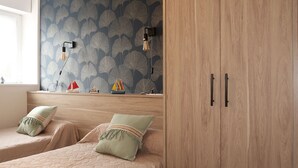 Une chambre avec deux lits, une armoire en bois, un mur orné d’un motif de feuilles et une étagère où sont exposés des modèles de bateaux.