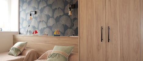 Un dormitorio con dos camas, un armario de madera, una pared con estampado de hojas y un estante con barcos modelo.