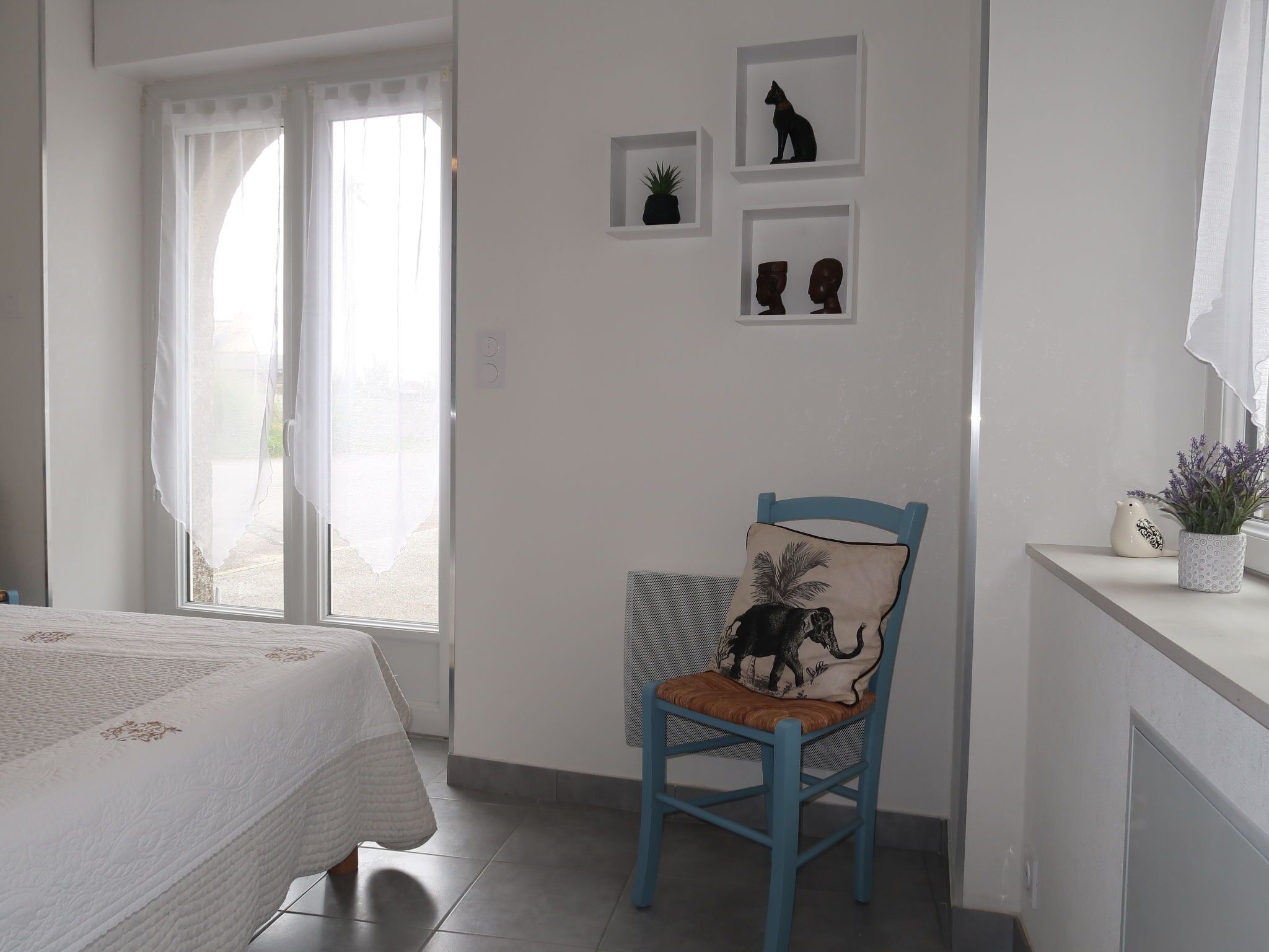 A bedroom with a bed, a chair with a cushion, a window with curtains, and wall-mounted shelves with decorative items.