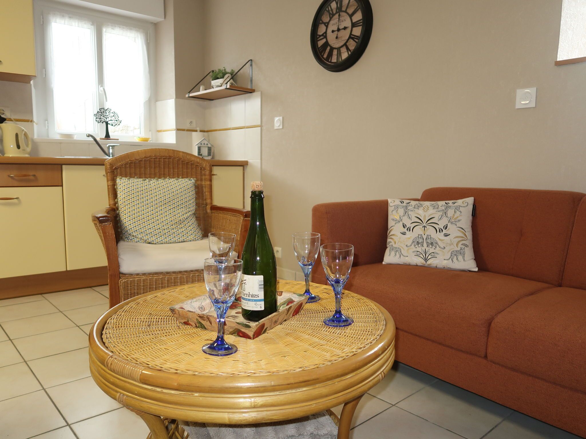 A living room with a round wicker table, a brown sofa, and a chair with a cushion. There is a bottle and glasses on the table.