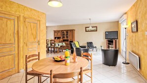 A dining area with a round wooden table, chairs, and a fruit bowl. There is a kitchen area with cabinets and a television in the background. - La Farniente by Interhome (La Turballe)