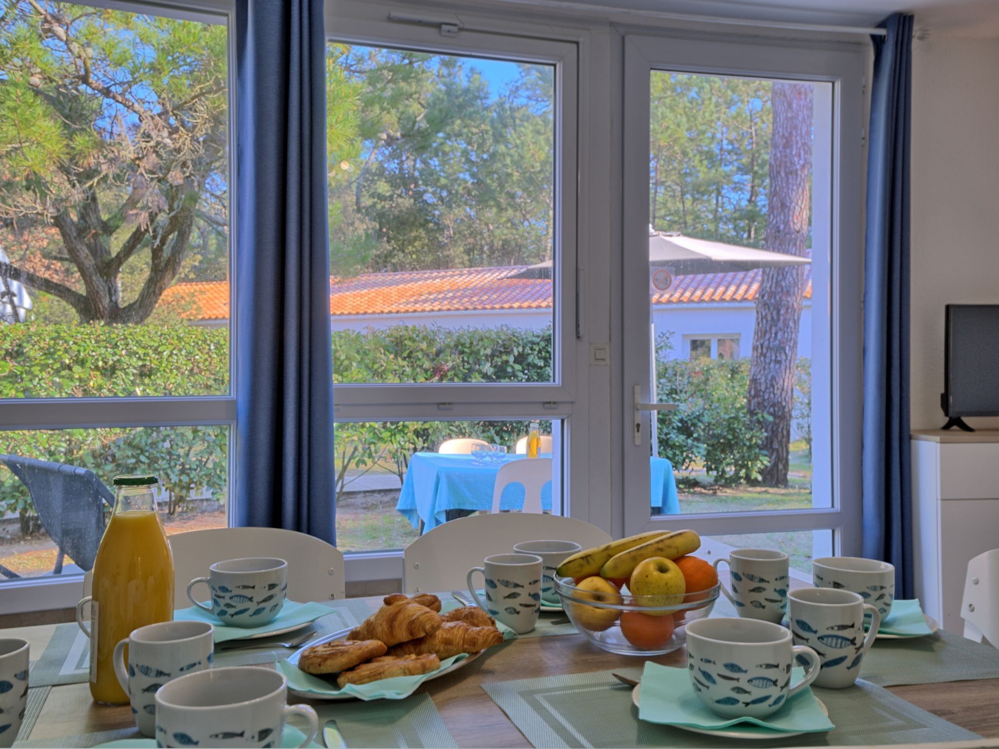 Et morgenbord med croissanter, frugt og kaffekopper, med udsigt til træer og bygninger gennem et vindue.
