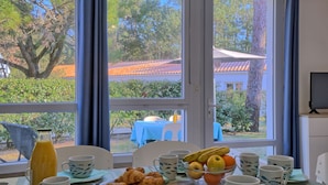 A breakfast table with croissants, fruit, and coffee cups, set against a window with a view of trees and buildings. - Hameau Océan by Interhome (Saint Hilaire de Riez)