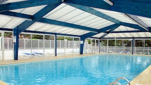 An indoor swimming pool with clear blue water, surrounded by white lounge chairs and a covered roof structure. - Hameau Océan by Interhome (Saint Hilaire de Riez)