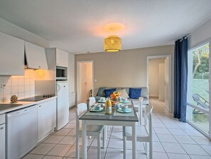 A modern kitchen with white cabinets, a dining area with a table set for two, and a view of the outdoors through large windows. - Hameau Océan by Interhome (Saint Hilaire de Riez)