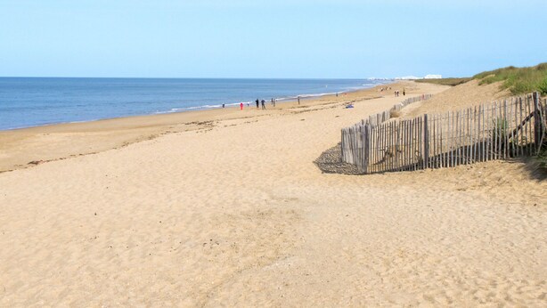 A wide sandy beach with a wooden fence on the right and a clear blue sky.