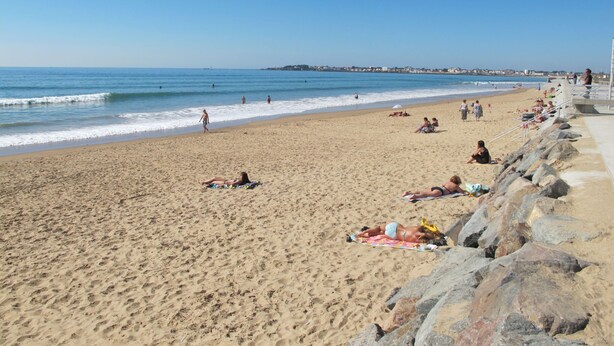 A sandy beach with people sunbathing and swimming, a rocky breakwater, and a clear blue sky.