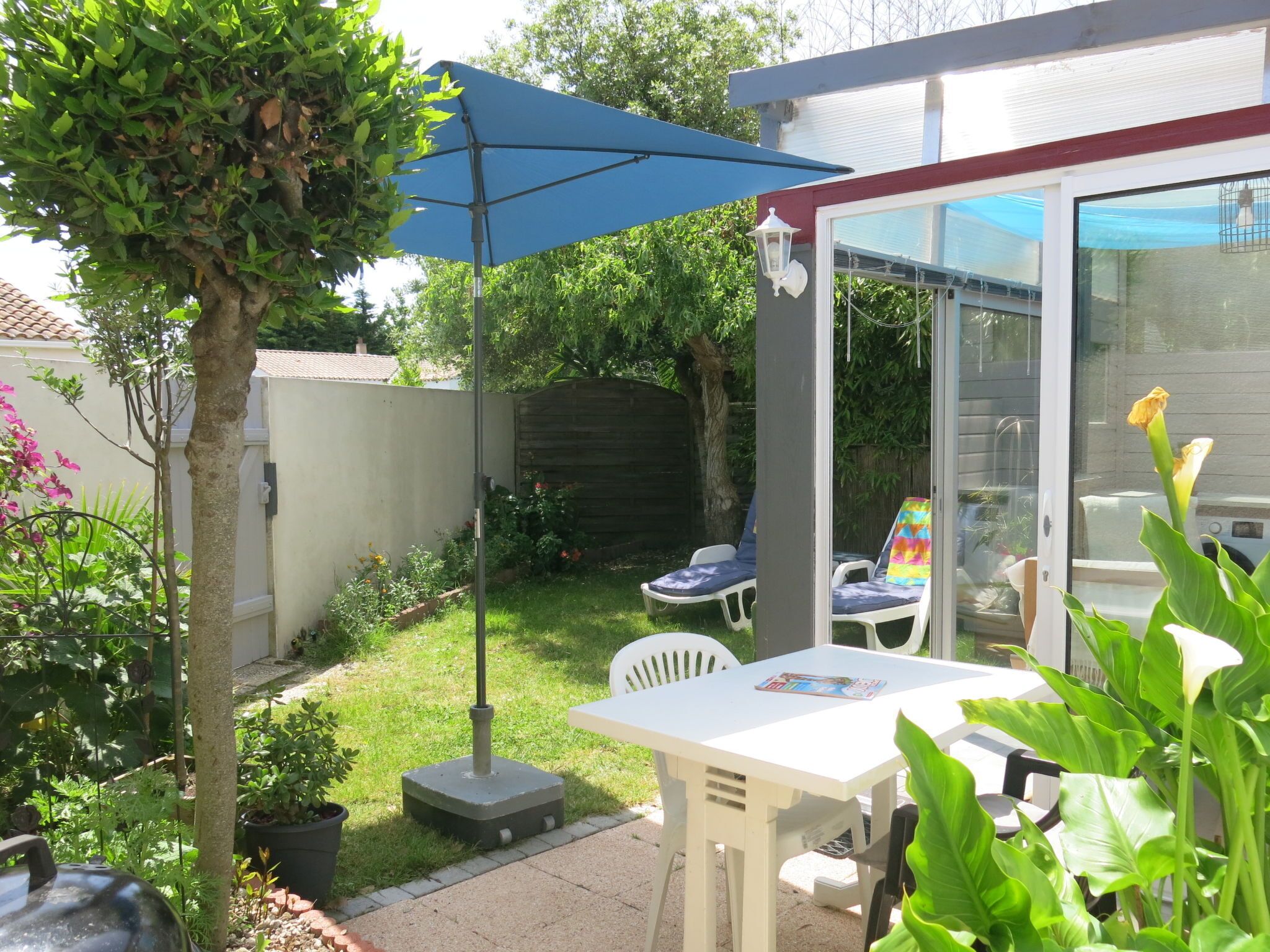 Eine Terrasse mit Tisch und Stühlen, einem blauen Sonnenschirm und einem Garten mit verschiedenen Pflanzen und einem Baum.