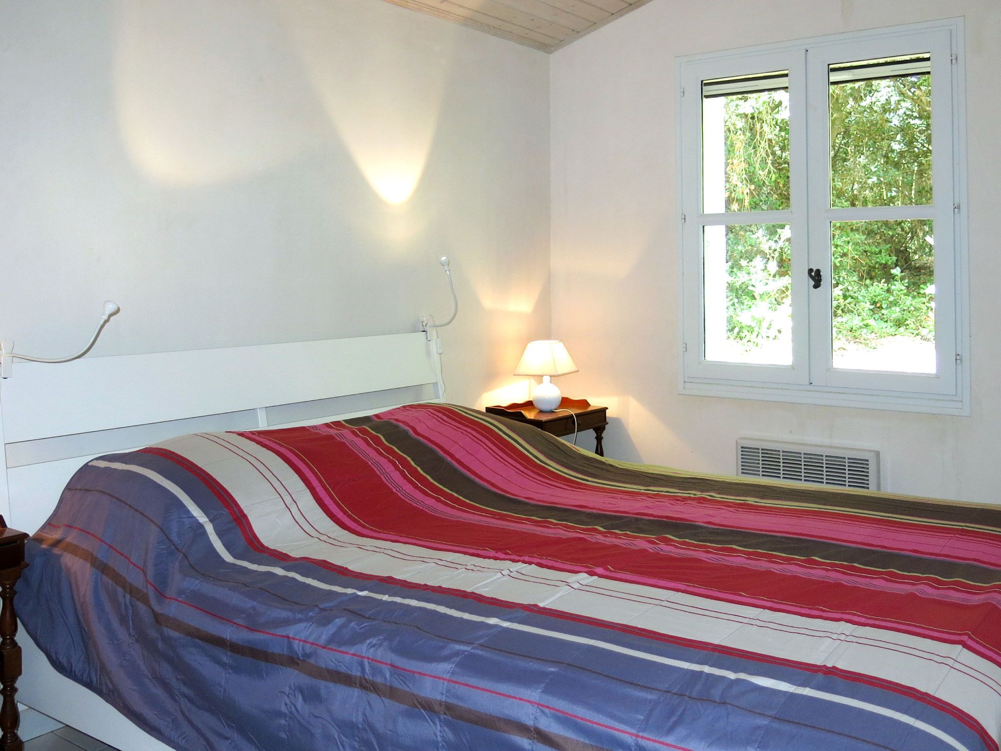 A bedroom with a bed, a bedside table with a lamp, a window with a view of greenery, and a wall socket.