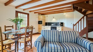 Una sala de estar acogedora con un sofá a rayas, una mesa de comedor de madera con sillas, una zona de cocina con electrodomésticos modernos y una escalera con barandillas de madera.