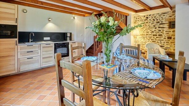 Un comedor acogedor con una mesa de madera dispuesta para cuatro personas, rodeada de sillas de mimbre. Un jarrón de cristal con flores está sobre la mesa, y se observa una pared de piedra al fondo.