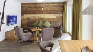 Un salon chaleureux avec un mur en bois, une tête de cerf accrochée au mur, un téléviseur, un canapé avec des coussins, deux fauteuils et une table basse en bois.