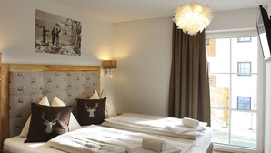 Une chambre d’hôtel avec un grand lit, deux oreillers ornés de motifs de cerfs, une tête de lit recouverte d’un tissu à motifs, une lampe fixée au mur et une fenêtre avec des rideaux.