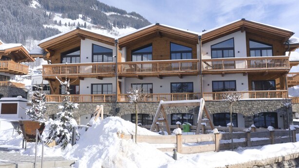 Un hôtel à plusieurs étages, doté de balcons en bois et de grandes fenêtres, qui se dresse sur fond de montagnes enneigées.