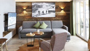 Un salon moderne avec un canapé gris, deux coussins verts, un téléviseur fixé au mur et une grande photo encadrée représentant des randonneurs en montagne.