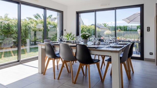 Un comedor moderno con una gran mesa dispuesta para seis personas, rodeado de amplios ventanales que ofrecen vistas a un patio con vegetación y una piscina.