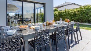 Un área de comedor al aire libre con una mesa y sillas modernas, un paraguas grande y vistas de la casa y el jardín.