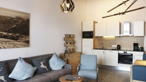 Uma sala de estar moderna com um sofá, mesa de centro e uma poltrona. Há uma área de cozinha com armários e eletrodomésticos.