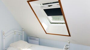 Un lit simple avec une tête de lit et un pied de lit blancs, une petite table de chevet et une fenêtre avec un cadre en bois.