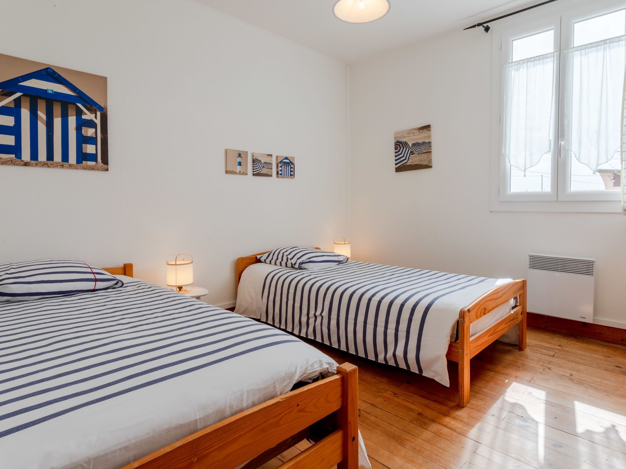Una habitación con dos camas de madera con ropa de cama a rayas, una mesita con una lámpara y un estante en la pared con objetos decorativos.
