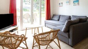 Un salon avec un canapé gris, des fauteuils en osier, un téléviseur à écran plat et une porte-fenêtre coulissante donnant sur un espace extérieur équipé d’une table et de chaises.