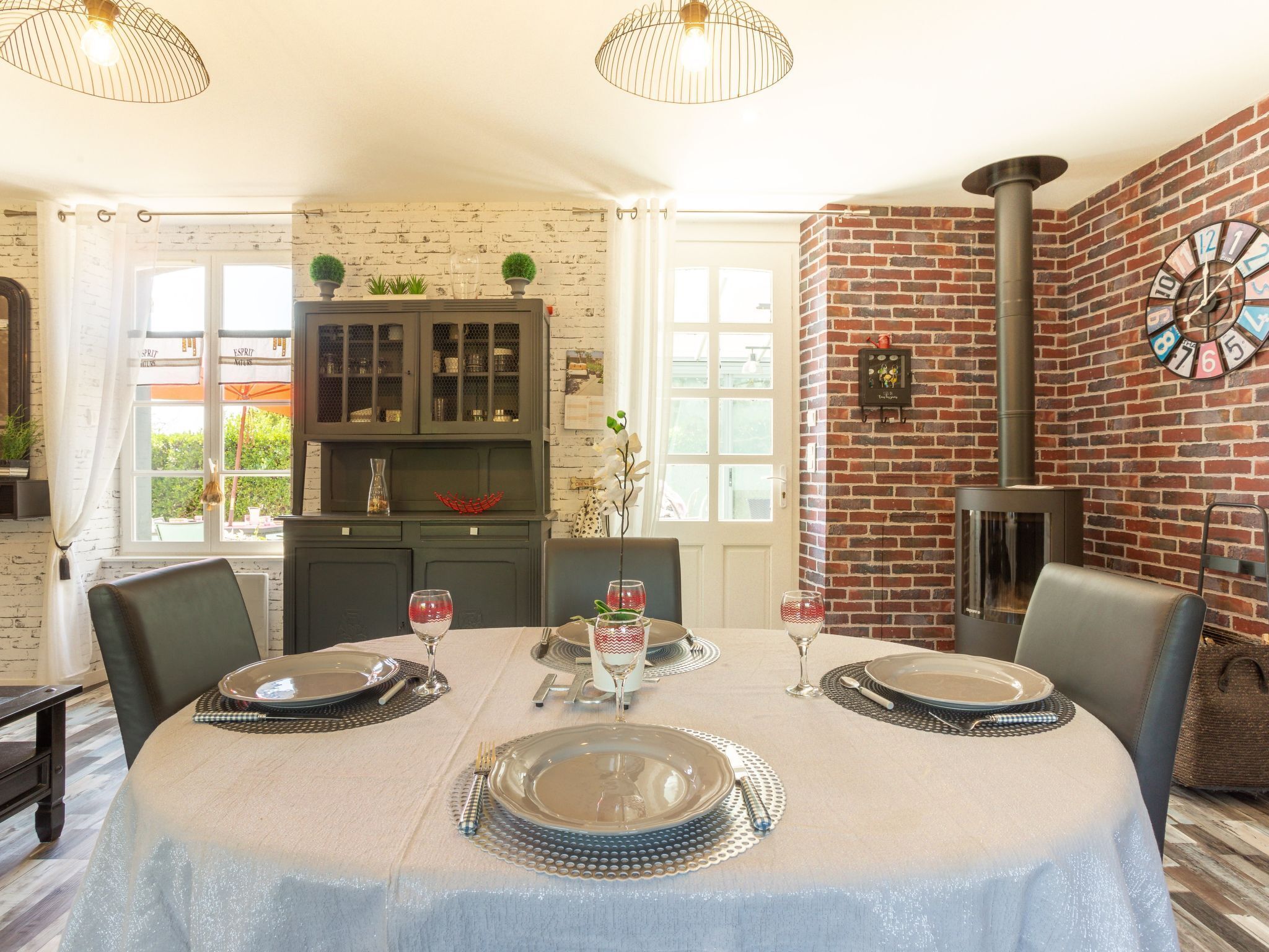 Un coin repas avec une table ronde dressée pour quatre personnes, un mur en briques orné d’une horloge et une cheminée.