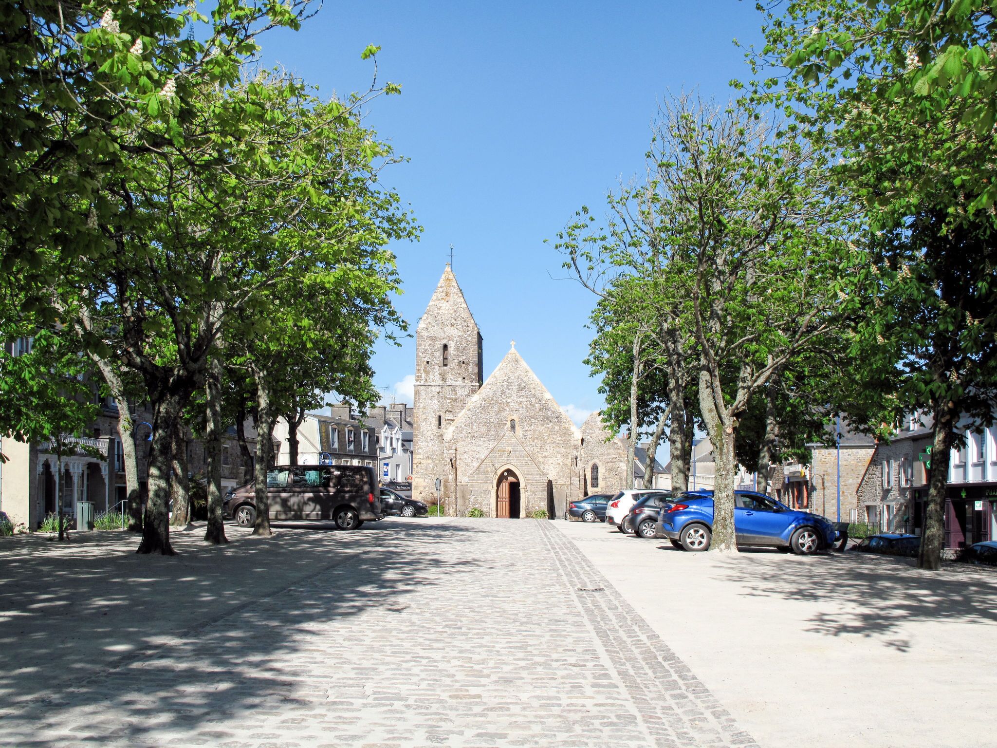 A cobblestone street leading to a historic stone church with a tall spire, surrounded by trees and parked cars.